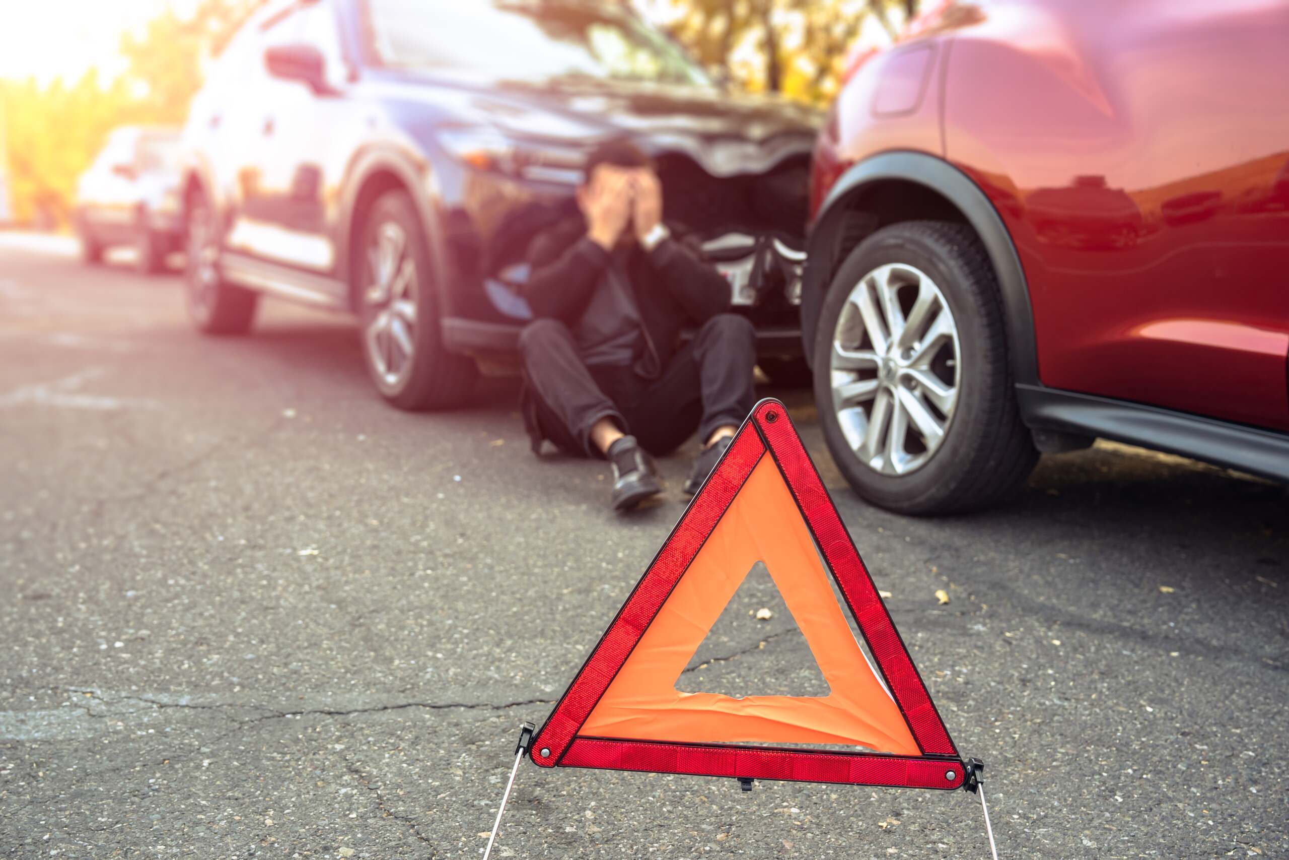 Distressed driver sitting on the road behind a red warning triangle after a rear end car collision.